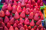 A large collection of bright red strawberries arranged in neat rows. The strawberries are fresh, glossy, and have green leaves at their tops, giving a vibrant and appetizing appearance.