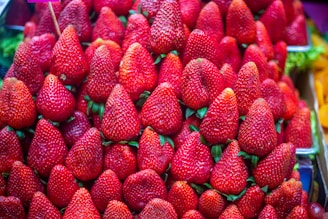 A large collection of bright red strawberries arranged in neat rows. The strawberries are fresh, glossy, and have green leaves at their tops, giving a vibrant and appetizing appearance.
