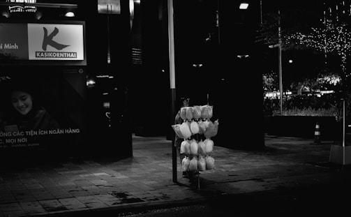 A dimly lit urban street scene at night features a stand with bags of cotton candy illuminated by nearby streetlights. In the background, there is a large advertisement with a smiling person on it. Sparse traffic cones and decorated trees with lights are visible, contributing to a quiet, nighttime atmosphere.