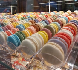 A colorful assortment of macarons is arranged in neat rows inside a display case. The macarons feature a variety of vibrant colors, including blue, pink, yellow, green, and more. They are presented in a way that showcases their smooth, round shapes and delicate textures.