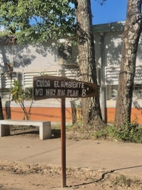 A wooden sign is displayed in front of a building and trees. The sign bears a message in Spanish urging environmental care. A white stone bench is situated nearby, with sunlight casting shadows from the trees around.