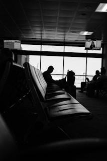 A dimly lit airport waiting area features rows of empty seats in the foreground, with silhouettes of travelers engrossed in their activities by the window. A departure sign and a TV screen are visible, adding to the typical airport ambiance. The windows provide a view of the tarmac outside.