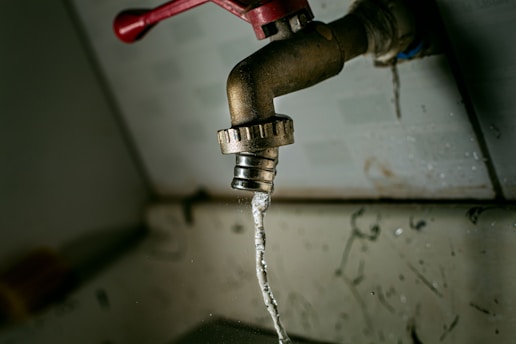 A close-up view of an old, slightly rusted faucet with water trickling out. The background is blurred, and there are visible water droplets scattered around. The red handle of the faucet provides a pop of color against the otherwise muted tones.