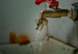 A rusty metal faucet with a red handle is mounted on a wall above a sink. Water is dripping from the faucet, emphasizing a worn or neglected appearance.