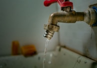 A rusty metal faucet with a red handle is mounted on a wall above a sink. Water is dripping from the faucet, emphasizing a worn or neglected appearance.