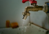 A rusty metal faucet with a red handle is mounted on a wall above a sink. Water is dripping from the faucet, emphasizing a worn or neglected appearance.
