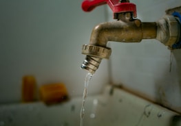 A rusty metal faucet with a red handle is mounted on a wall above a sink. Water is dripping from the faucet, emphasizing a worn or neglected appearance.
