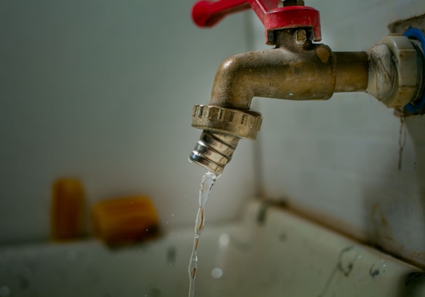 A rusty metal faucet with a red handle is mounted on a wall above a sink. Water is dripping from the faucet, emphasizing a worn or neglected appearance.