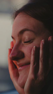 Softly lit close-up of a smiling woman gently touching her glowing skin in a bright, airy room.