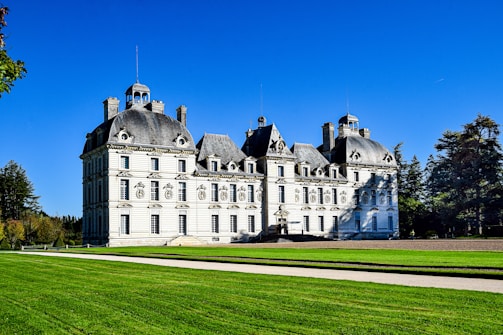 A grand, multi-story château with ornate architectural details and steep slate roofs set against a bright blue sky. A manicured lawn and trees surround the building, highlighting its elegance and symmetry.