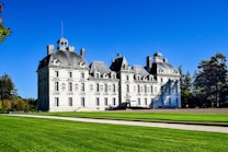 A grand, multi-story château with ornate architectural details and steep slate roofs set against a bright blue sky. A manicured lawn and trees surround the building, highlighting its elegance and symmetry.