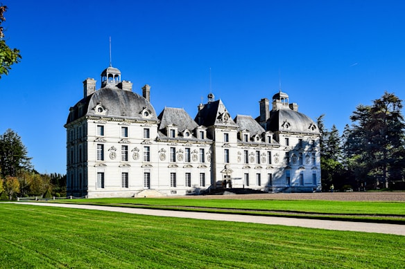 A grand, multi-story ch&acirc;teau with ornate architectural details and steep slate roofs set against a bright blue sky. A manicured lawn and trees surround the building, highlighting its elegance and symmetry.
