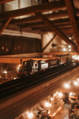A miniature model train is displayed on elevated tracks within a warmly lit, wooden interior space. The train is detailed with various cars, and small lights create a cozy atmosphere. The background shows structured wooden beams adding a rustic feel.