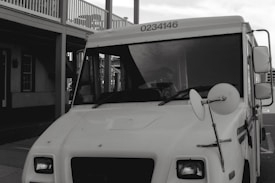 A white delivery truck with the number 0234146 displayed at the top, parked next to a building with a porch and multiple windows. A side mirror is prominently visible on the truck.