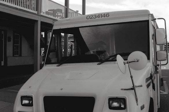 A white delivery truck with the number 0234146 displayed at the top, parked next to a building with a porch and multiple windows. A side mirror is prominently visible on the truck.