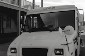 A white delivery truck with the number 0234146 displayed at the top, parked next to a building with a porch and multiple windows. A side mirror is prominently visible on the truck.