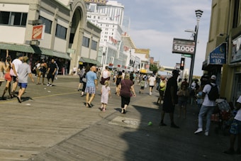 A busy boardwalk scene with a variety of people walking and interacting. Shops with colorful signs and advertisements line the sides of the boardwalk, suggesting a lively commercial area. The sky is clear, adding to the bright daytime setting.