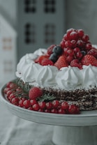 A beautifully decorated cake topped with an assortment of fresh berries, including strawberries, blueberries, raspberries, and currants. The cake is adorned with white whipped cream swirls and sits on an elegant cake stand. Sprigs of evergreen add a festive touch.
