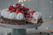 A beautifully decorated cake is topped with an assortment of fresh berries, including strawberries and redcurrants. It features layers of cream and chocolate shavings on the sides, with dollops of whipped cream around the top edge. A slice has been removed, revealing the intricate layers inside. The background is softly lit with twinkling lights, enhancing the festive atmosphere.