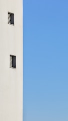 A minimalist image featuring a tall, white wall on the left side with small, rectangular windows evenly spaced. The rest of the space is dominated by a clear blue sky.