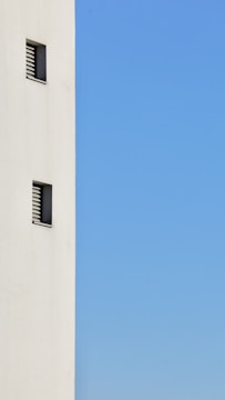 A minimalist image featuring a tall, white wall on the left side with small, rectangular windows evenly spaced. The rest of the space is dominated by a clear blue sky.