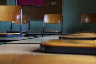 Several wooden chairs with metal legs are arranged inside a classroom. The focus is on the seat backs, with desks in the foreground slightly out of focus. A bulletin board is visible in the background.