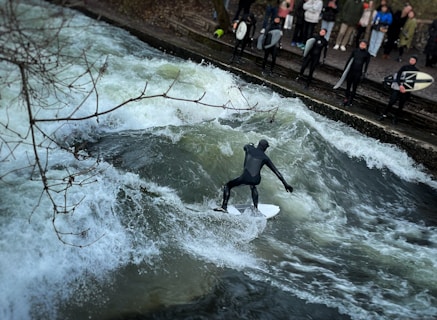 A person in a wetsuit is surfing on a river wave, surrounded by spectators watching from the riverbank. The scene is dynamic with splashing water and the surfer maintaining balance on the board. Several people in similar attire are gathered, some holding surfboards, while leafless branches partially frame the view.