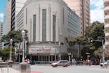A historic building with an art deco design features ornate vertical windows and geometric patterns. The entrance sign reads 'cine theatre Brasil Vallourec.' The street in front has a few cars and a group of people. Lamp posts and trees line the sidewalk, and other tall buildings are in the background.