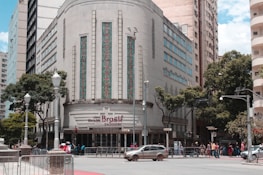 A historic building with an art deco design features ornate vertical windows and geometric patterns. The entrance sign reads 'cine theatre Brasil Vallourec.' The street in front has a few cars and a group of people. Lamp posts and trees line the sidewalk, and other tall buildings are in the background.