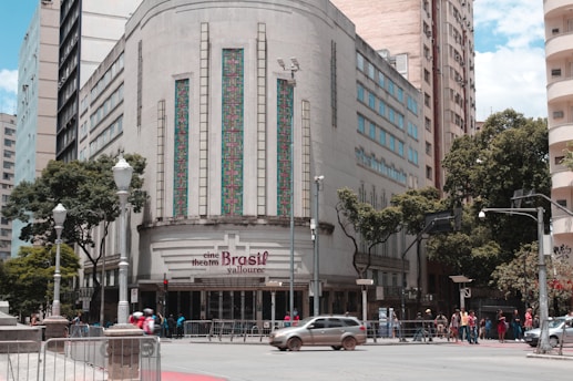 A historic building with an art deco design features ornate vertical windows and geometric patterns. The entrance sign reads 'cine theatre Brasil Vallourec.' The street in front has a few cars and a group of people. Lamp posts and trees line the sidewalk, and other tall buildings are in the background.