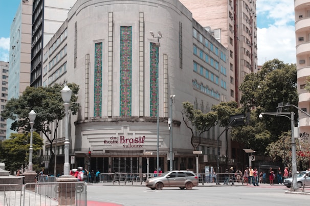 A historic building with an art deco design features ornate vertical windows and geometric patterns. The entrance sign reads 'cine theatre Brasil Vallourec.' The street in front has a few cars and a group of people. Lamp posts and trees line the sidewalk, and other tall buildings are in the background.