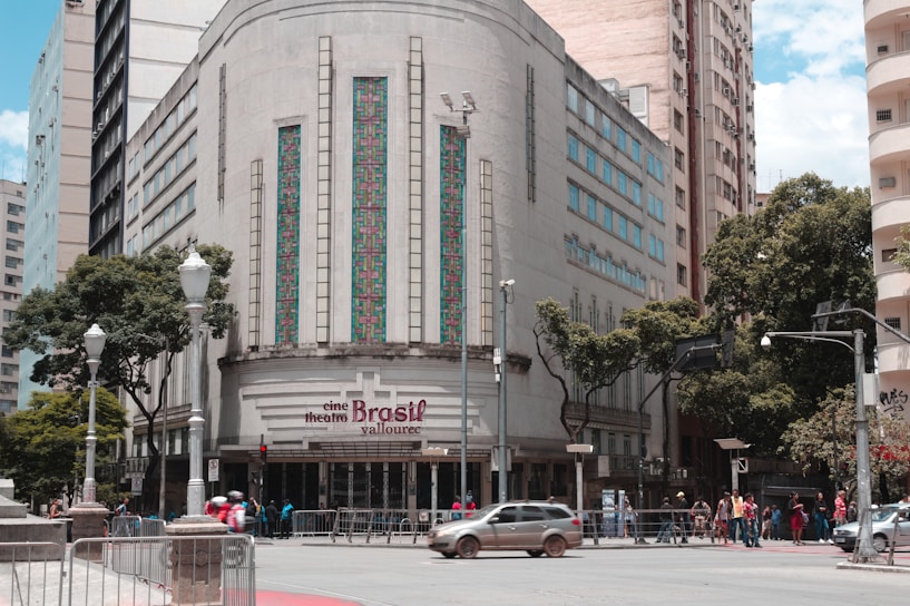 A historic building with an art deco design features ornate vertical windows and geometric patterns. The entrance sign reads 'cine theatre Brasil Vallourec.' The street in front has a few cars and a group of people. Lamp posts and trees line the sidewalk, and other tall buildings are in the background.