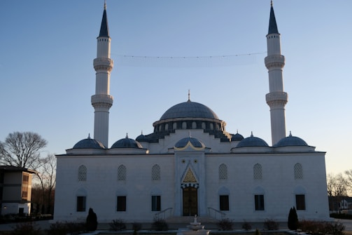 A mosque with two tall minarets and a large central dome, surrounded by smaller domes. The building has a symmetrical, elegant design with light-colored walls and intricate window details. It's set against a clear sky with minimal surrounding trees and structures.