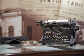 A vintage typewriter sits on a reflective glass surface, accompanied by a pair of round spectacles with brown frames. Behind them, there's a blurred background with sepia-toned newspaper text, contributing to an antique and historical ambiance.