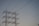 A series of high-voltage power lines are suspended on a metal lattice tower, silhouetted against a clear sky. The structure features multiple crossbeams and insulators, emphasizing the complex engineering involved in electricity transmission.