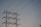 A series of high-voltage power lines are suspended on a metal lattice tower, silhouetted against a clear sky. The structure features multiple crossbeams and insulators, emphasizing the complex engineering involved in electricity transmission.
