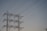 A series of high-voltage power lines are suspended on a metal lattice tower, silhouetted against a clear sky. The structure features multiple crossbeams and insulators, emphasizing the complex engineering involved in electricity transmission.