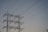 A series of high-voltage power lines are suspended on a metal lattice tower, silhouetted against a clear sky. The structure features multiple crossbeams and insulators, emphasizing the complex engineering involved in electricity transmission.