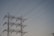 A series of high-voltage power lines are suspended on a metal lattice tower, silhouetted against a clear sky. The structure features multiple crossbeams and insulators, emphasizing the complex engineering involved in electricity transmission.