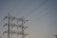 A series of high-voltage power lines are suspended on a metal lattice tower, silhouetted against a clear sky. The structure features multiple crossbeams and insulators, emphasizing the complex engineering involved in electricity transmission.
