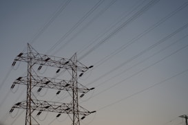 A series of high-voltage power lines are suspended on a metal lattice tower, silhouetted against a clear sky. The structure features multiple crossbeams and insulators, emphasizing the complex engineering involved in electricity transmission.