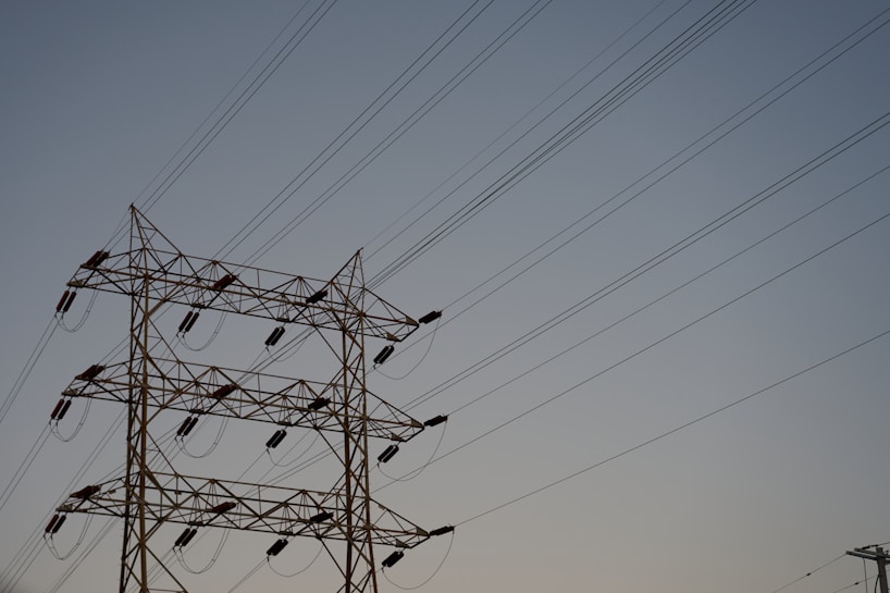 A series of high-voltage power lines are suspended on a metal lattice tower, silhouetted against a clear sky. The structure features multiple crossbeams and insulators, emphasizing the complex engineering involved in electricity transmission.