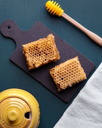 Two pieces of honeycomb sit on a black slate cutting board. A wooden honey dipper with a yellow top is placed above the board. Nearby, a yellow ceramic honey pot is partially visible, placed on a teal-colored surface alongside a light blue cloth.