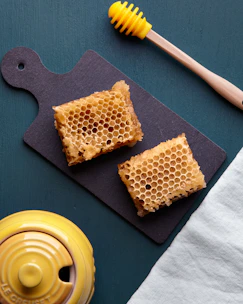 A silicone brush resting on a wooden cutting board next to a jar of honey and fresh herbs.