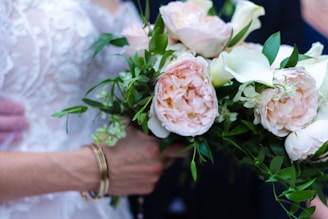 A close-up of a person holding a bouquet of flowers which includes pale pink roses and white lilies, surrounded by green foliage. The person is wearing an intricately designed white lace garment and a gold bracelet on their wrist.