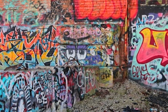 An urban scene featuring a wall covered in vibrant graffiti artwork, with a variety of colors and abstract designs. The wall's surface is mostly brick, with some concrete sections. The ground is littered with debris, adding to a sense of urban decay.