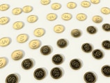 A scattered pattern of gold and dark coins with dollar signs embossed on them, set against a light background.