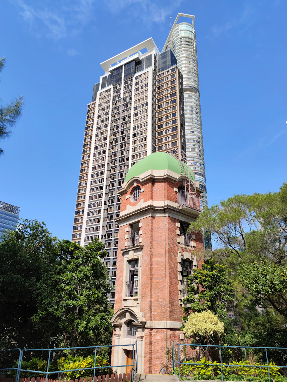 The image depicts a tall, modern skyscraper with a green dome on its top and a smaller, older brick building with a clock tower at its base. The two structures are situated next to each other, creating an interesting juxtaposition between old and new architectural styles. In the foreground, there is a lush park filled with trees, providing a natural contrast to the urban landscape.