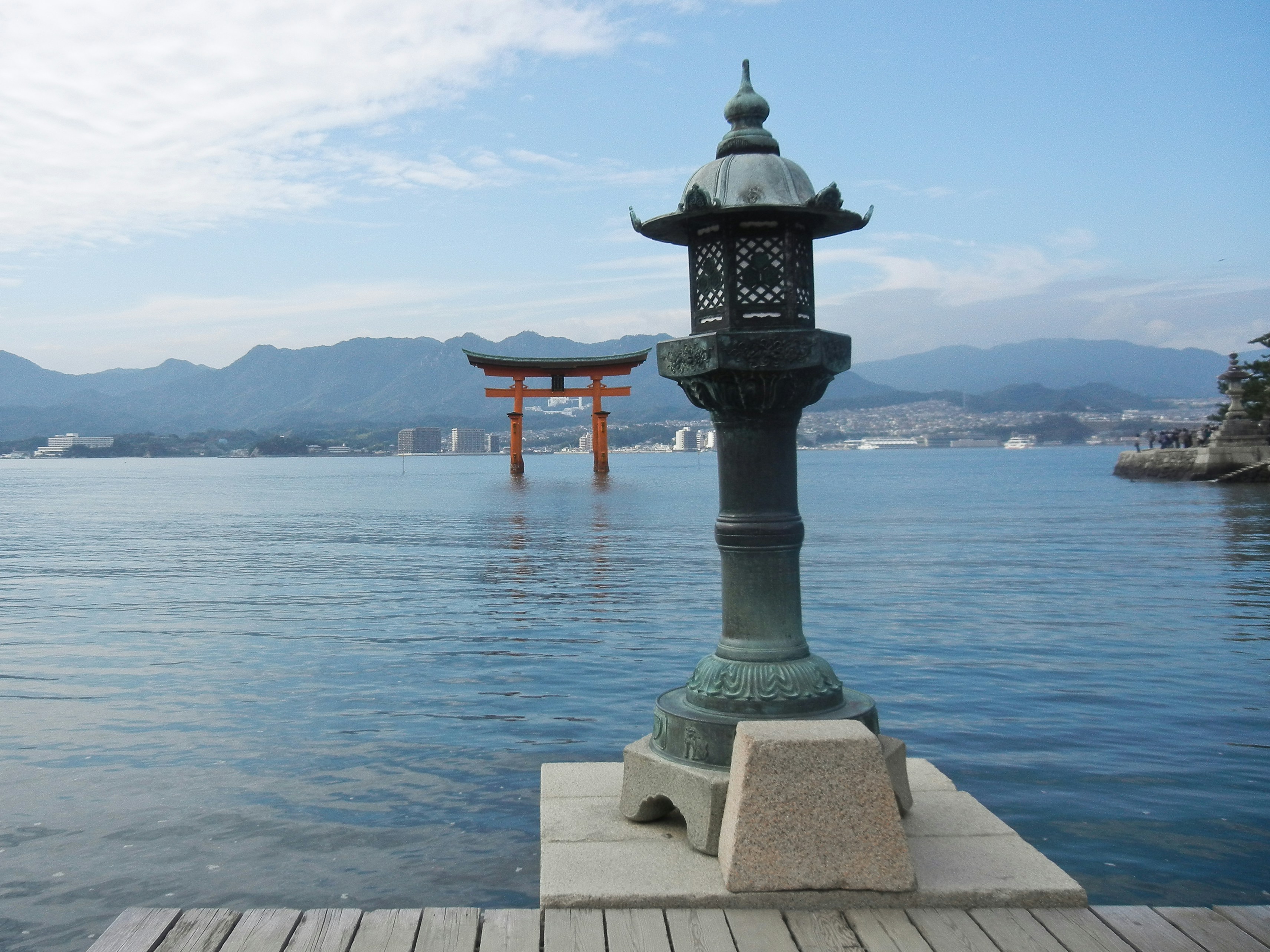 Stone lantern on a concrete jetty overlooks calm water with a distant red torii gate and hazy mountains.