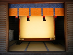 A traditional Japanese room with tatami mat flooring and a wooden frame at the center. The frame is adorned with intricate golden designs at the corners. Above it is a decorative hanging with a blue and orange pattern featuring tassels in black and red.